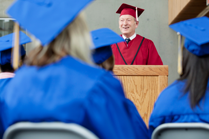 Older man in graduation robes speaking to a graduating class